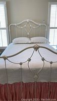 Full view of the antique iron full bed frame with bedspread, sheets, and pillows, showing white painted metal with floral decorative accents on headboard and footboard.