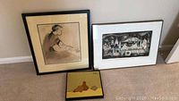 Three framed artworks displayed on the floor against the wall, including an ink and watercolor drawing, a pears canvas artwork, and a black and white signed print.