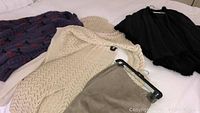Four garments on a bed including black knit sweater with buttons, cream knit sweater, black wool cape with fur trim, and beige suede skirt.