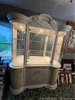 Front view of large ornate display cabinet with glass doors and lighting showing three shelves inside and lower storage compartments.