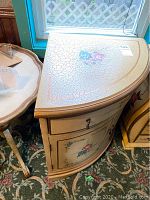 Front and side angle view of wooden corner table with floral designs on top, drawer, and cabinet doors.