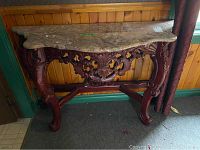 Full view of side table showing marble top and carved wooden base near a wooden wall and green doorframe.