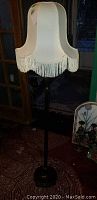 Full view of floor lamp showing cream fabric shade with fringe and dark brown base with small feet.