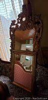 Tall ornate mirror with carved floral crest leaning against furniture near window