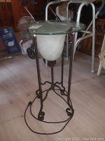 Full view of the urn style table lamp showing the clear round glass top, white urn-shaped glass lamp body, and black metal stand with decorative curved elements and a visible power cord.