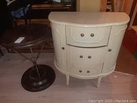Photo showing white half-moon shaped dresser with three drawers and side compartments with doors, next to a round dark wood top occasional table with metal frame and base, floor visible.