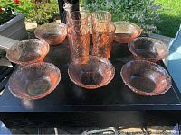 Seven pink glass soup bowls arranged in a semicircle with four pink drinking glasses centered behind them on a black surface outdoors.
