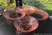 Four pink glass serving pieces arranged on a black surface outdoors, showing two round pie-plate-style bowls, one smaller round bowl elevated, and one oblong bowl.