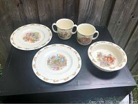 Five ceramic Bunnykins pieces: two 8-inch plates, porringer bowl, one two-handled mug, one one-handled mug, all displayed on a black surface outdoors.