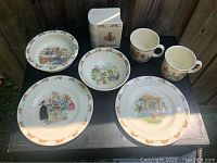 Overview photo showing all seven items: two plates, two mugs, two bowls and one baby bank. Items arranged on black surface with wooden fence background.