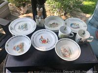 Full set of Wedgwood Peter Rabbit dishes including plates, bowls, mugs, and a miniature stuffed Peter Rabbit figure displayed on a table.