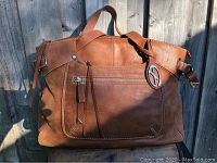 Front full view showing large tan leather purse with front zip pocket, thick stitched straps, and metal Francesco Biagia emblem charm.