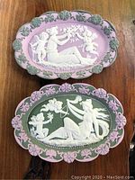 Two oval-shaped antique Jasperware plaques placed on a wood surface showing full view of both plaques with detailed relief work of cherubs and women.