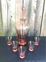 Front view of the pink glass decanter with five shot glasses arranged around it, showing the height and shape of the decanter and glasses.