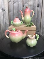 Four piece apple motif ceramic tea set includes teapot, coffee pot, covered sugar bowl and creamer on a black surface with burlap box background