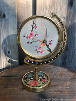 Full view of Chinese silk screen art in brass and enamel rotating frame on wooden surface with outdoor background.