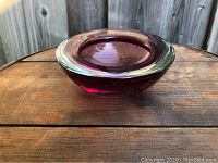 Side angle view of a small heavy glass bowl with deep purple and clear glass layers, placed on a wooden surface with a background of wooden fencing.