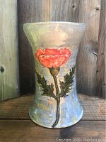 Full view of ceramic vase showing raised red poppy flower with green leaves on muted gray-blue background, standing on wood surface with wood panel background.