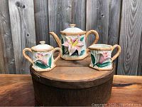 Full view of three-piece Fitz and Floyd tea set on rustic wooden surface, showing the teapot, creamer, and sugar bowl with tiger lily floral motifs.