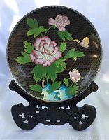 Front view of vintage cloisonne brass plate on carved wooden stand showing floral and butterfly enamel design on black background.