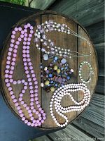 Five pieces of pink jewelry displayed on wooden surface: four necklaces and one bracelet with various size beads and colors including light pink, white, and pastel tones.