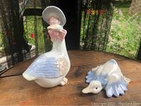 Mother Goose figurine standing beside the gosling figurine on a wooden surface with mesh screen and garden background.