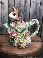 Full side view of ceramic bunny teapot resting on wooden surface showing the large bunny lid and floral decorations.