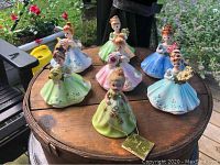 Seven pastel-colored porcelain birthday doll figurines arranged on a wooden table outdoors, each holding flowers and wearing detailed skirts.