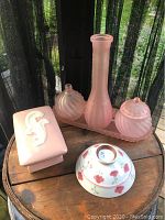 Six decorative pink items arranged on a wooden surface including three glass pieces, a ceramic bowl, and two covered containers.