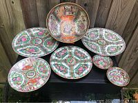 Seven pieces of vintage Chinese Famille Rose porcelain dishes displayed outdoors on a black surface with a wooden fence background, showing front and interior designs including a large bowl and assorted plates.