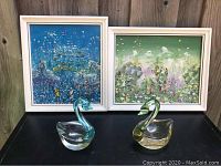 Photo showing two framed paintings with botanical and bird themes alongside two glass swan figurines in blue and green tints.