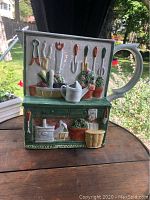 Front view showing garden tools, plants, and watering can design on garden shed shaped ceramic piece.