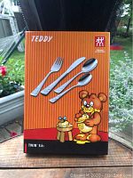 Front view of the Henkel Teddy child cutlery set box showing four pieces: fork, knife, tablespoon, teaspoon, with teddy bear image and branding on orange striped background.