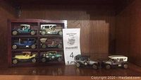 Nine miniature vintage cars arranged on a wooden shelf along with a small wooden display case. The cars have detailed wheels and convertible tops.