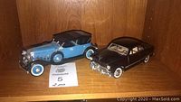 Photo showing two model cars on a wooden shelf: a blue and black vintage convertible and a maroon sedan.