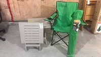 Two green deluxe foldable lawn chairs with one chair folded and standing upright, accompanied by folded beige plastic side tables with slatted tops.