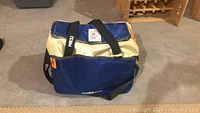 Front angled view of blue and beige Mobicool cooler bag with black shoulder strap on concrete floor near wooden shelf.