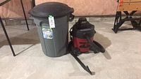 Photo showing gray plastic garbage can with black lid next to small red and black Shop Vac on concrete floor basement.