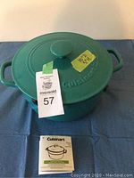 Green enameled cast iron pot with lid on blue fabric, Cuisinart manual below the pot, lot tag visible.