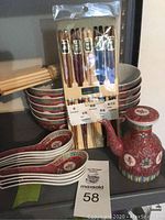 Asian soup set displayed on shelf including 10 patterned bowls stacked, red and patterned soup spoons, packaged lacquered chopsticks, bundled wooden chopsticks, and a decorative teapot.