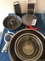Photo showing various kitchen items on a blue surface. Visible items include metal mixing bowls, metal bundt pan, two metal box graters, red measuring cups, white plastic strainers, and a collapsible metal steamer basket.