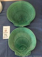Two green glaze scallop shell plates displayed on blue surface with a marked auction tag.