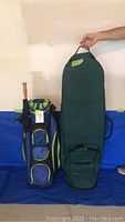 Blue and black golf carry bag and green padded golf travel bag side by side against wall.