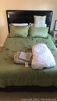 Queen size bed with green comforter embroidered with floral design, 5 pillows including 2 green large pillows, 1 decorative floral pillow, and 2 white pillows. Green bedding set, black bed skirt visible.