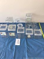 All nine square glass canisters arranged on a blue cloth, showing their lids and seals, alongside the three glass mugs