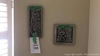 Two rectangular and two square wall hangings with indigenous style black designs on silver backgrounds, mounted on a wall.