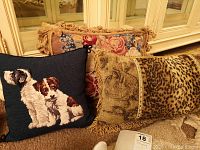 Three throw cushions displayed together on a carpet in front of a mirrored cabinet: black embroidered dog cushion, leopard print cushion with fringe, and floral embroidered cushion with tassels.