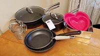 Wide view showing black speckled non-stick pots and pans with lids, metal square baking pan, pink heart-shaped plastic bake tins and two clear glass measuring cups on wooden table.