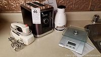 Photo showing all four items together on a kitchen counter: hand mixer, toaster, food chopper, and kitchen scale.