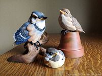 Three Kaiser bird figurines including Blue Jay on log, Baby Songbirds Sparrow on pot, and smaller Baby Songbirds resting figurine on wood table surface.